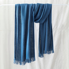 Soft blue banana yarn scarf with fringe detailing draped over a white clothing rack against a neutral linen backdrop.
