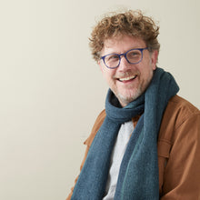 Smiling man wearing a soft blue woven banana yarn scarf with casual winter clothing in a minimalist indoor setting.