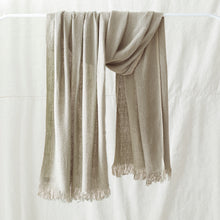 Soft natural beige banana yarn scarf with fringe detailing draped over a white clothing rack against a neutral linen backdrop.