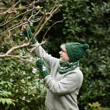 Man wearing forest green cable knit wool bobble hat with matching wool wristwarmers and neckwarmer outdoors in winter garden