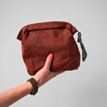 Hand holding a rust red woven cotton pouch bag with decorative fabric zip pull against a clean neutral background.