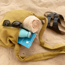 Mustard yellow cotton crossbody bag on sand with beach essentials including sunglasses, towel, book and water bottle.