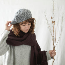 Woman wearing a grey cable knit hat and soft plum banana yarn scarf with neutral winter clothing in a minimalist studio setting.